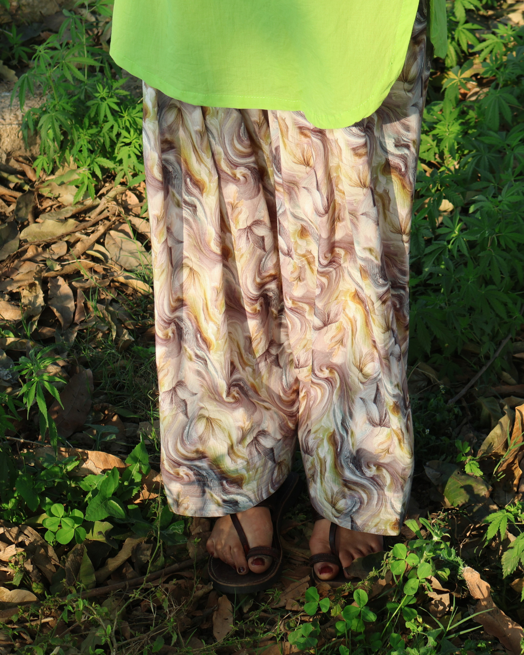 Person wearing a green top and patterned pants standing in a natural setting with greenery.