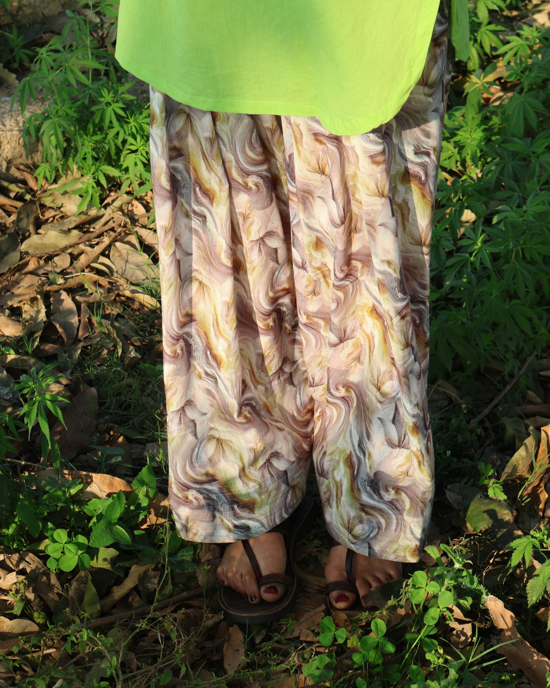 Person wearing a green top and patterned pants standing in a natural setting with greenery.