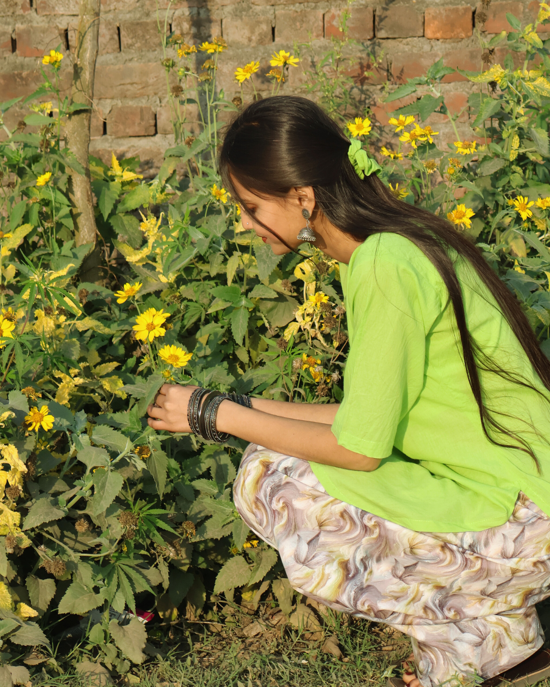 Woman in a green shirt and patterned pants interacting with yellow flowers in a garden.
