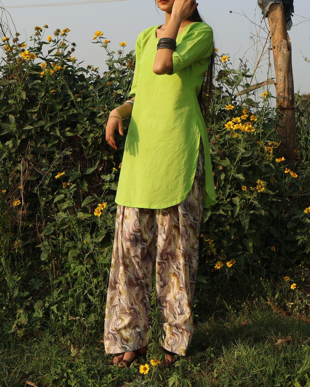 Person wearing a bright green top and patterned pants standing in a natural setting with plants and flowers.