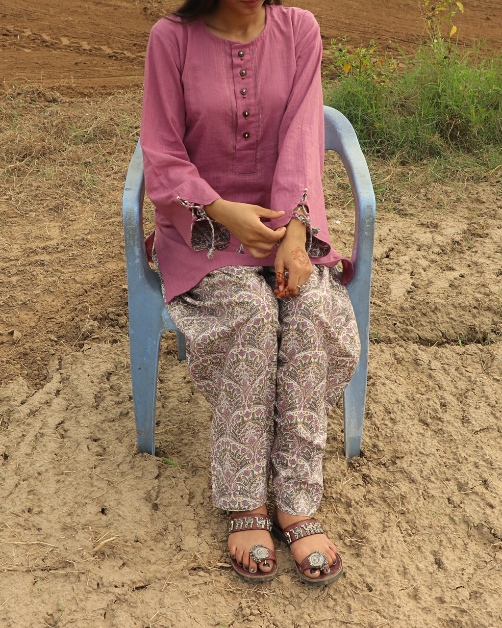 A woman wearing a pink long-sleeve kurta and patterned trousers sits on a light blue plastic chair outdoors on sandy ground.