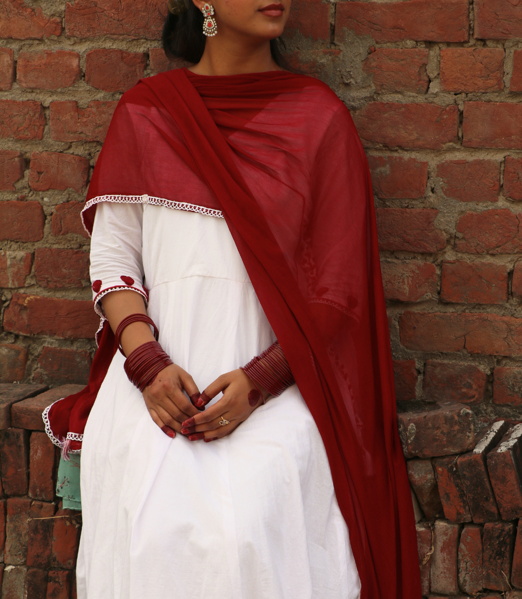 Woman wearing a white dress with a red dupatta against a brick wall.