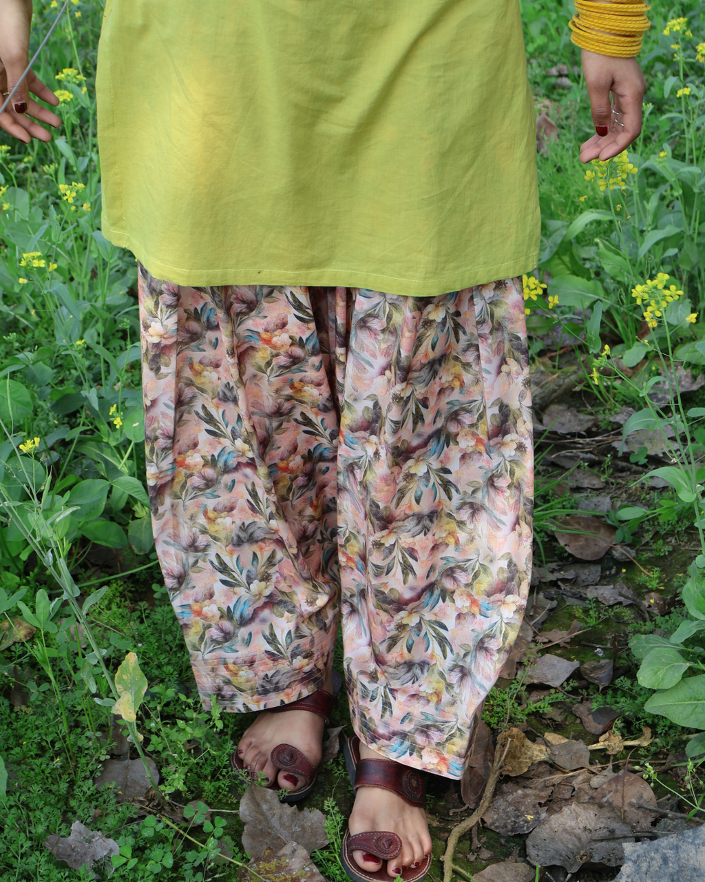 Person wearing a green top and floral pants standing in a field with yellow flowers.