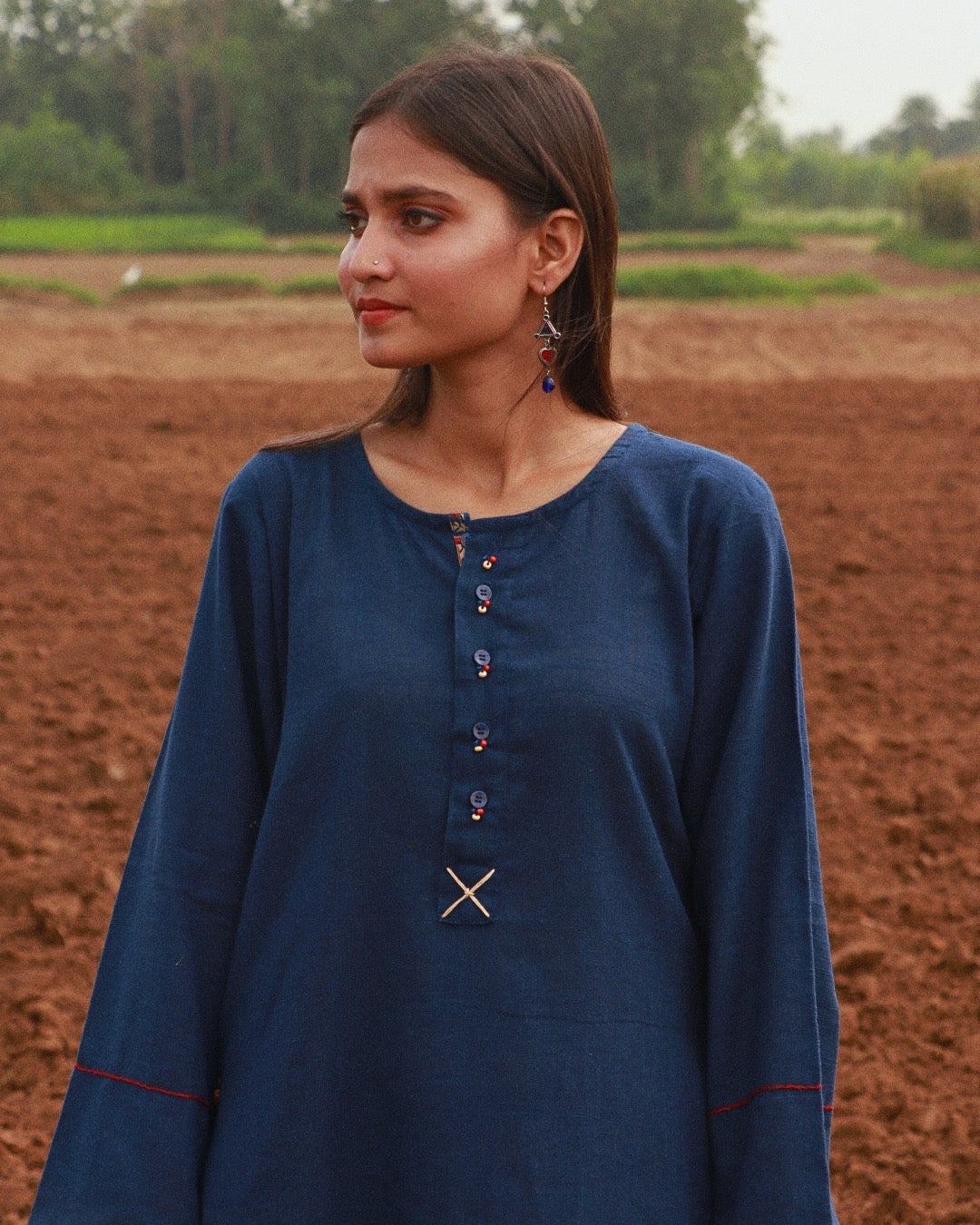 A woman wearing a dark blue long-sleeve tunic top with decorative buttons and a small embroidered detail at the neckline, standing in an outdoor field of reddish-brown soil.