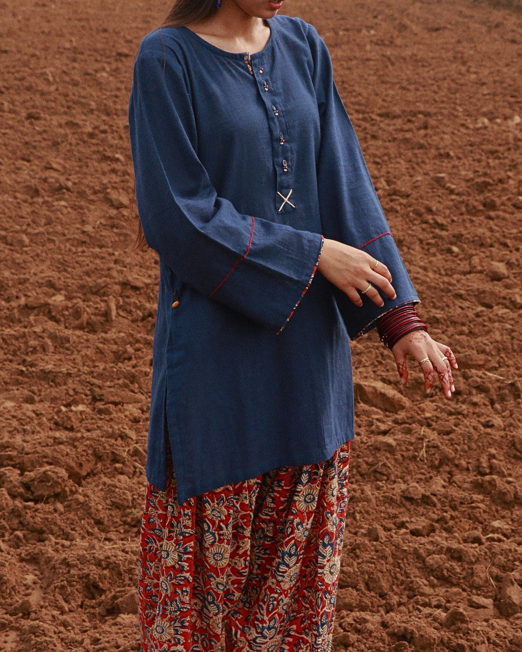 A woman wearing a blue long-sleeve kurta with red contrast stitching and a red, blue, and white patterned skirt, standing in front of a brown dirt background.