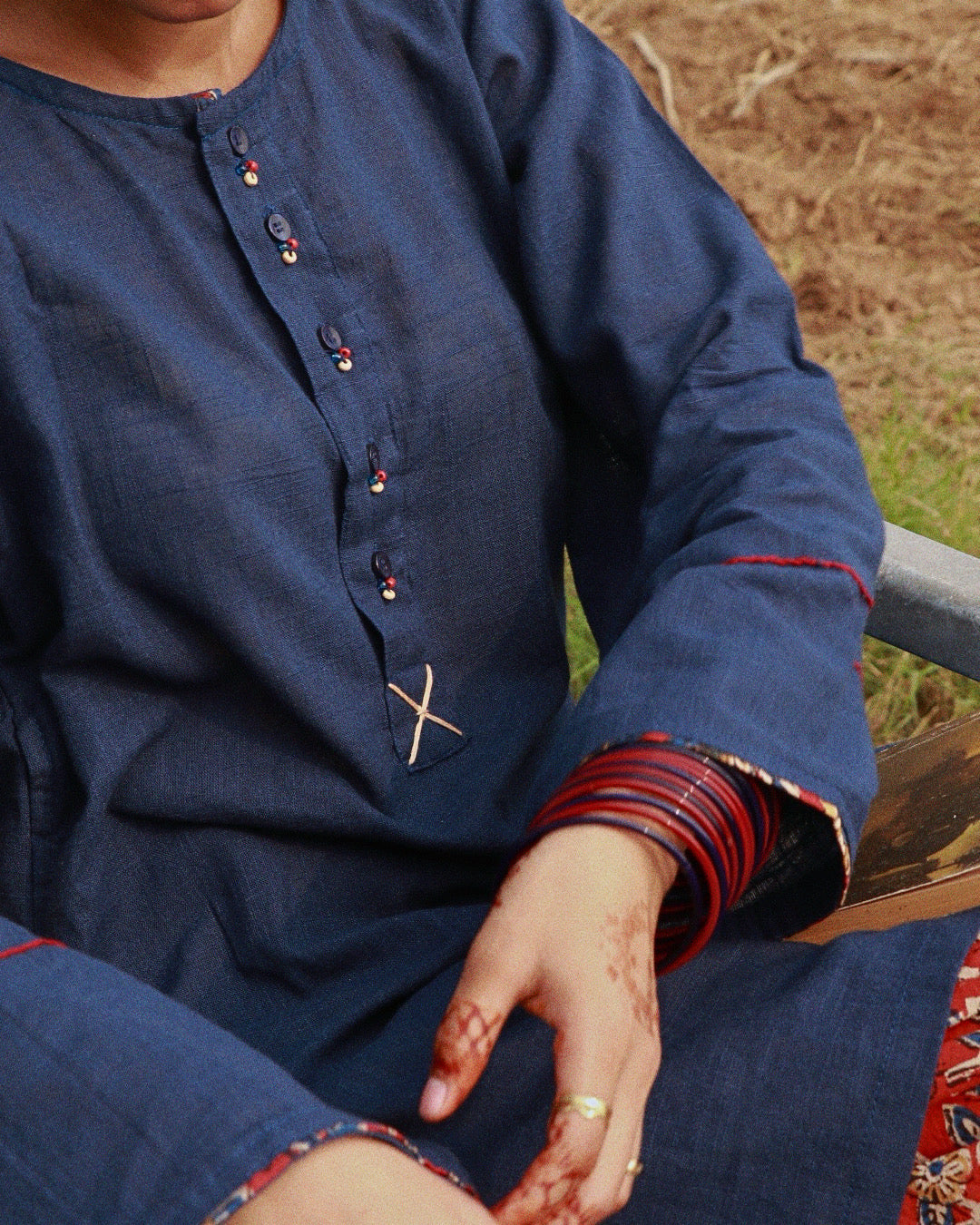 A close-up of a person wearing a dark blue long-sleeve kurta with decorative embroidered details, red striped cuffs, and a small white cross motif on the chest. The person is seated outdoors with dry grass and a chair visible in the background.