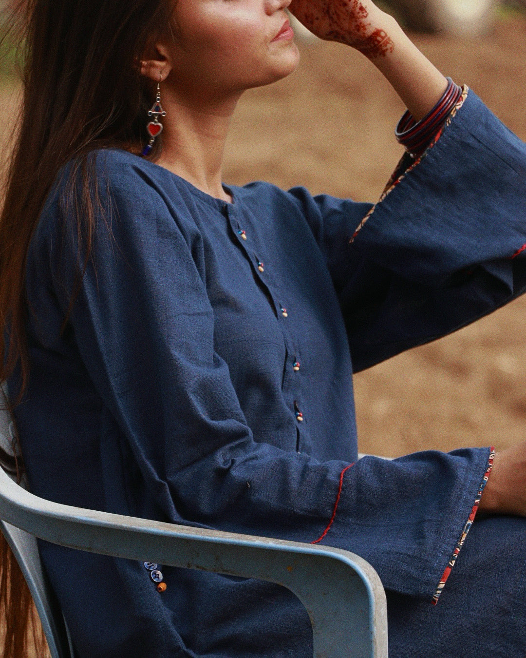 A woman wearing a dark blue long-sleeve tunic with colorful embroidered trim on the cuffs and button placket, seated in a light blue chair against a blurred outdoor background.