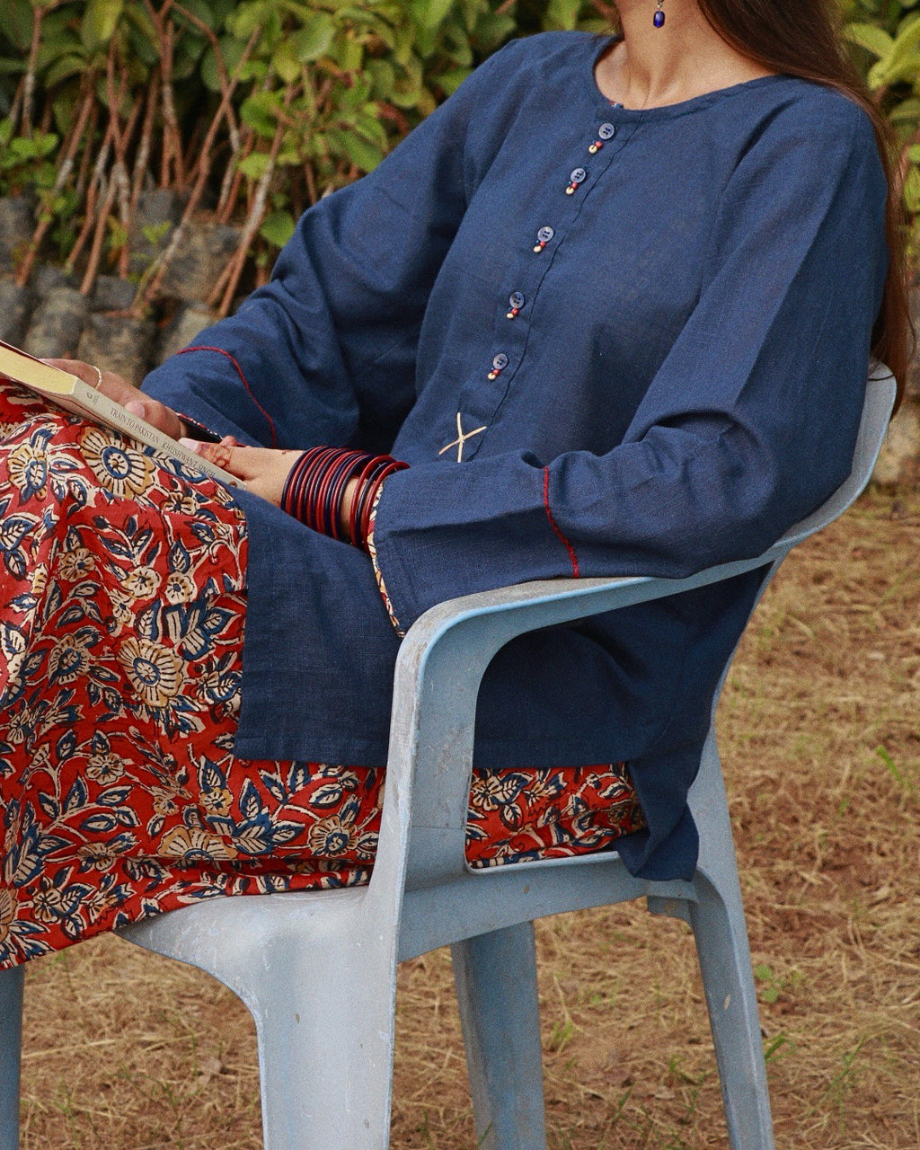 A woman seated outdoors on a light blue plastic chair, wearing a navy blue long-sleeve tunic top with decorative buttons and a red floral printed skirt. She is holding a book, with foliage and dry grass in the background.