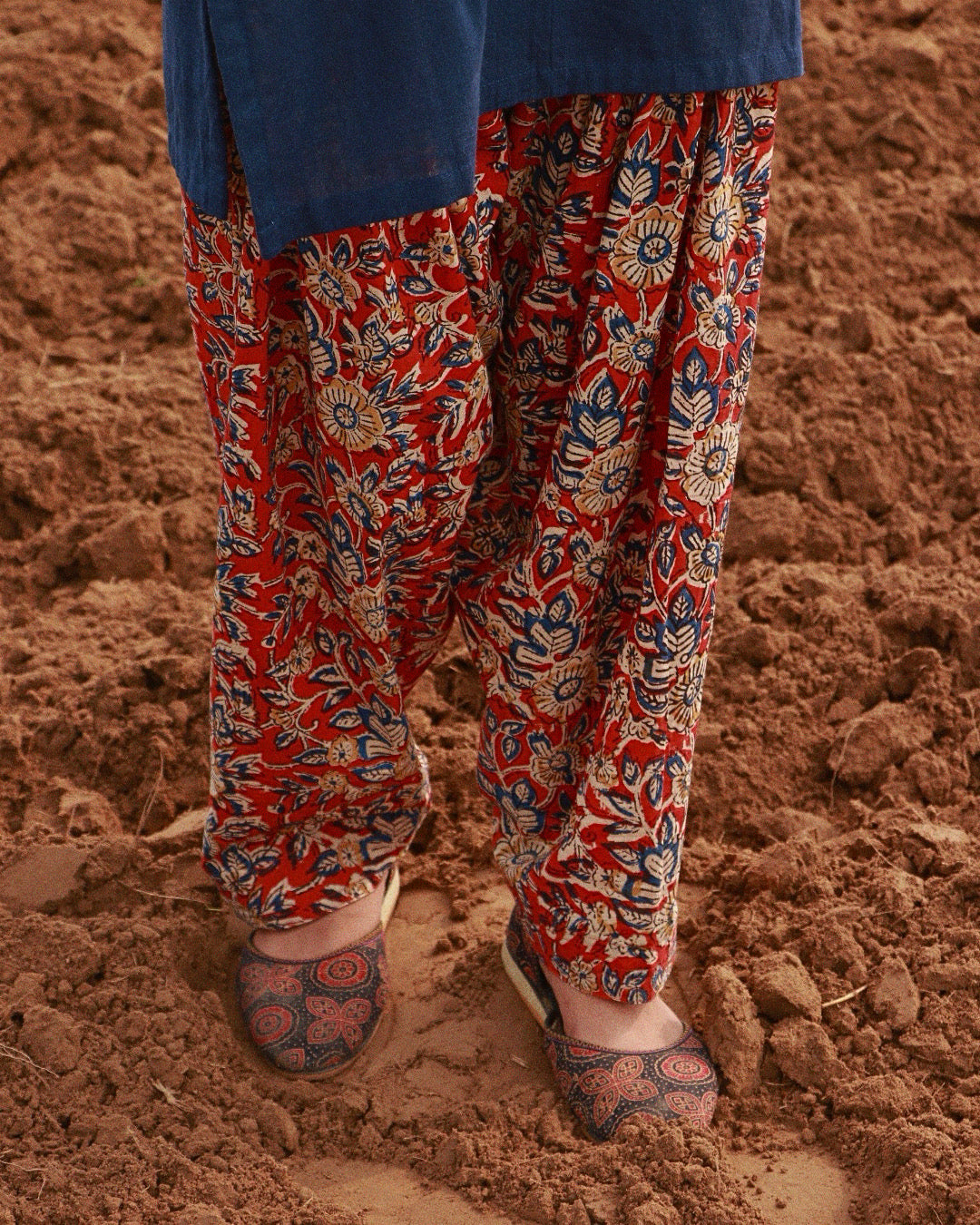 A person wearing red patola-style pants with a blue and beige floral block print, standing in loose brown soil. A blue top is partially visible at the top edge, and patterned flat shoes are shown at the feet.