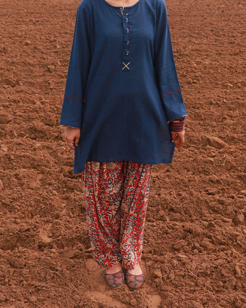 A person wearing a dark blue long-sleeve tunic with decorative buttons and red piping, paired with red and white patterned pants, standing on reddish-brown soil.