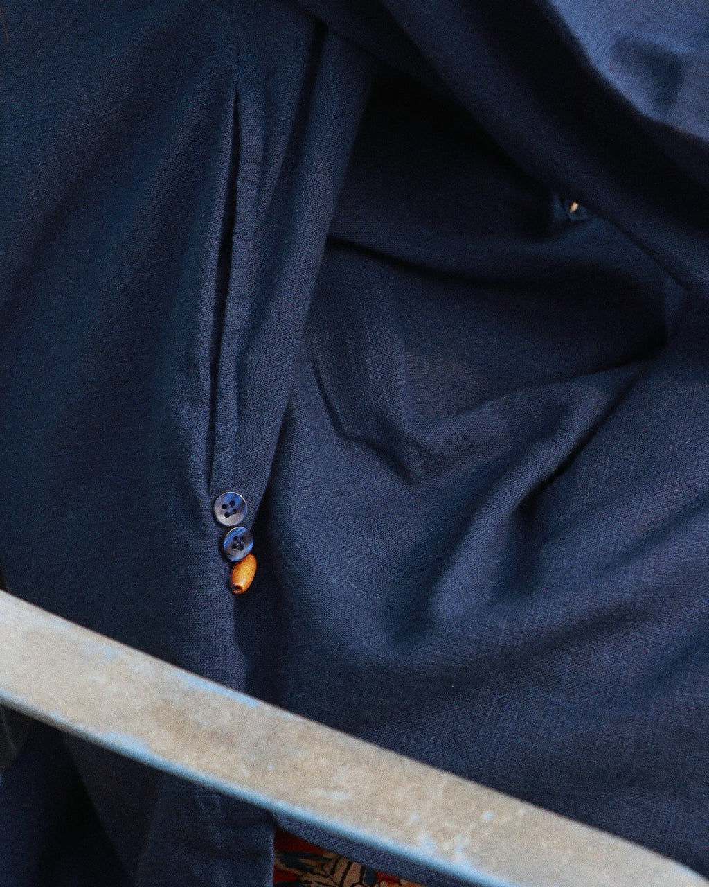 Close-up of a dark navy blue fabric garment with a buttoned pocket and a small amber bead accent, shown in a detailed product shot.