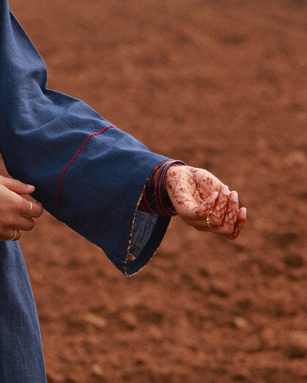 A close-up of a person's hand and wrist wearing a dark blue sleeve with red contrast stitching, set against a blurred reddish-brown background.