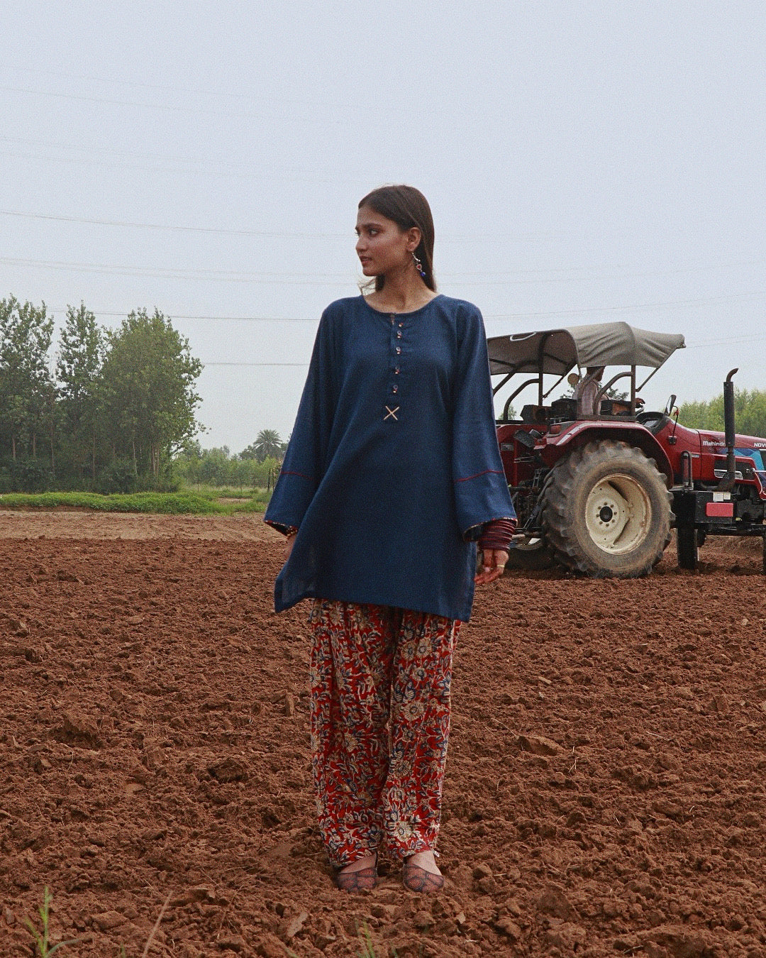 A woman stands in a plowed field wearing a navy blue tunic with a small cross detail at the neckline and red floral patterned pants. A red tractor is visible in the background under an overcast sky.