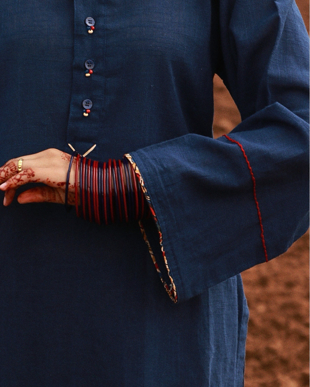Close-up of a person wearing a dark navy blue kurta with wide flared sleeves, red and gold embroidered trim at the cuffs, and stacked red bangles on one hand.