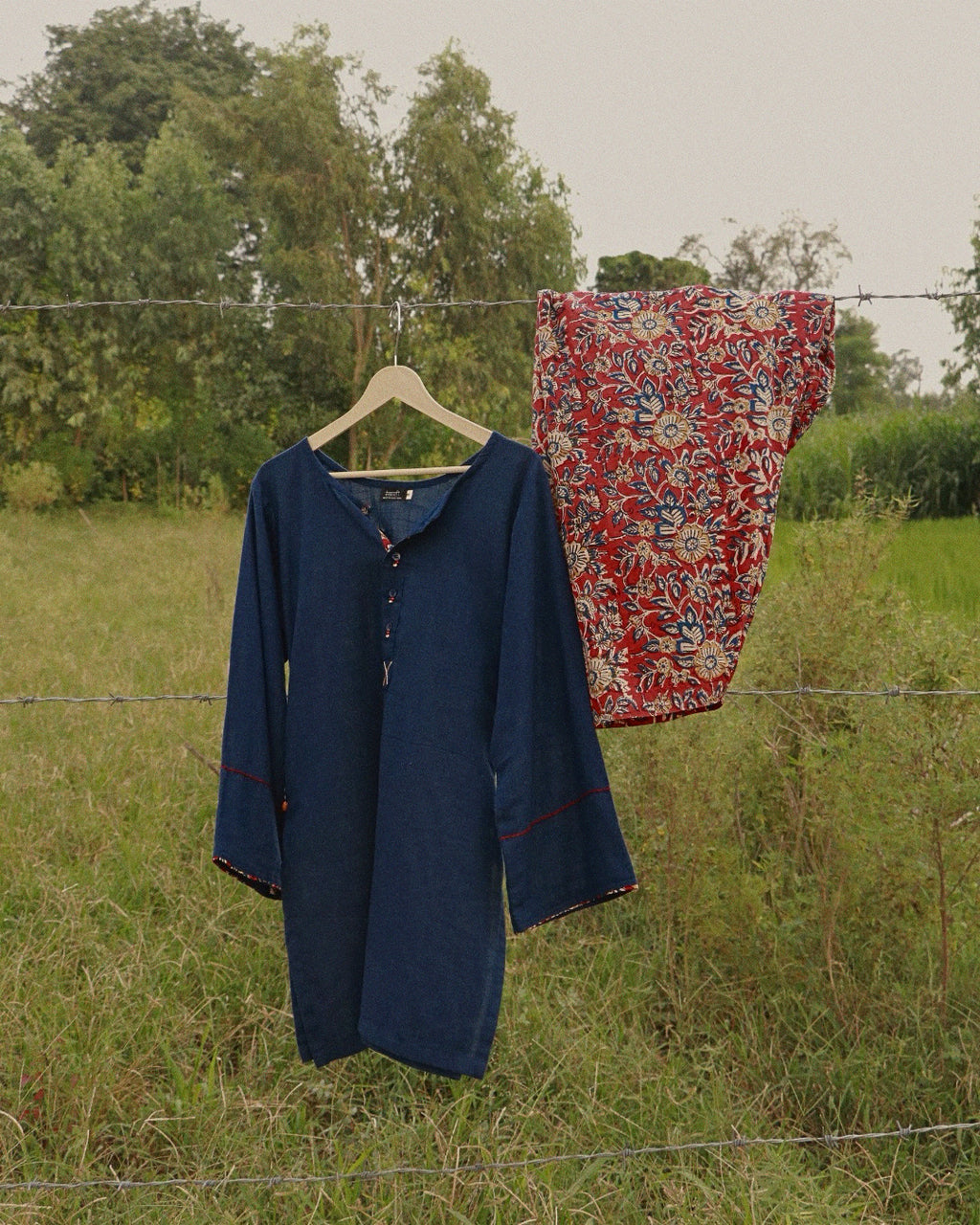 A navy blue long-sleeve tunic with red piping details hangs on a wooden hanger from a wire fence in an outdoor grassy field, next to a red and beige patterned fabric piece.